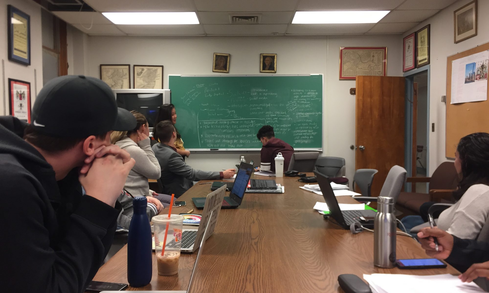A group of students sits around a long conference table with laptops, facing a green chalkboard filled with writing; one student stands near the board