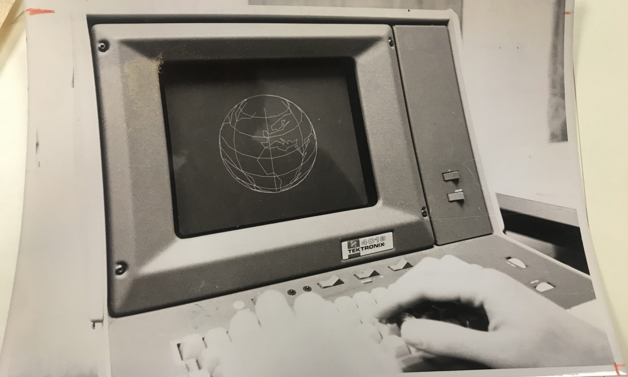 Black and white photo of a retro computer terminal displaying a wireframe globe, with a hand typing on the keyboard.